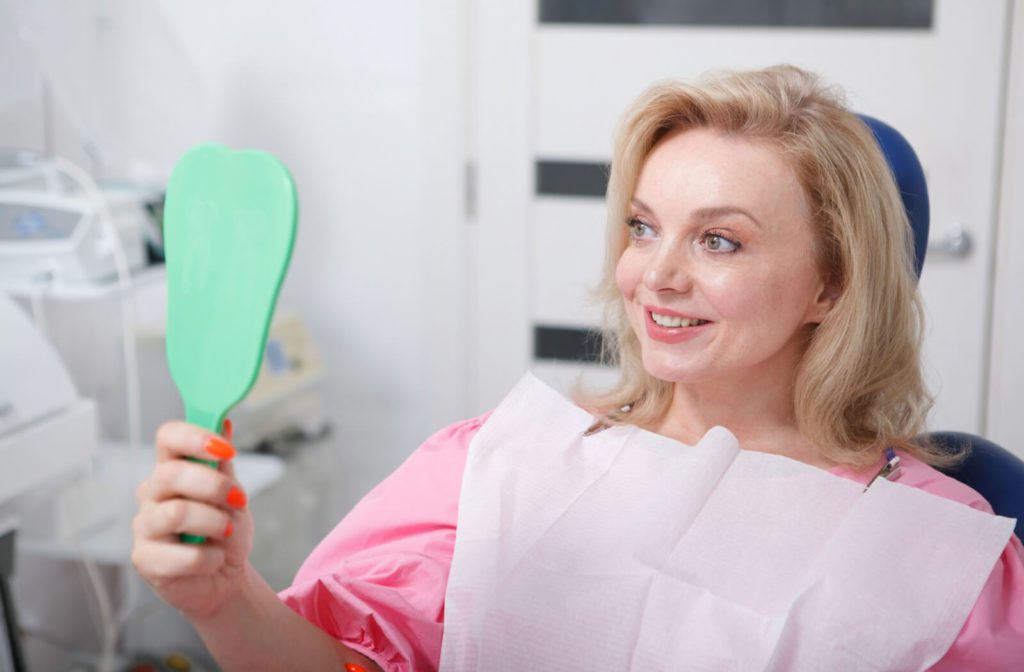 A woman with a dental bib sitting in a dentist’s chair and smiling into a handheld mirror